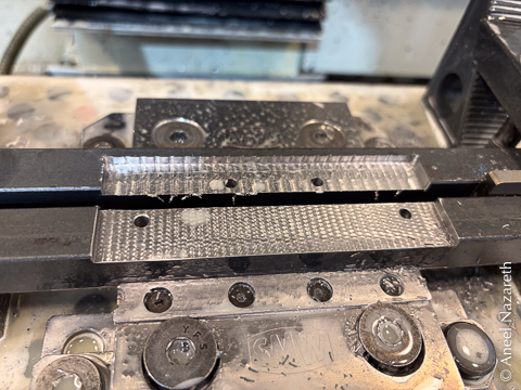 Two bars are held in a vise with a spacer between them. A rectangular recess has been milled that spans both bars, but doesn't extend all the way through. There are also screw holes in an arc pattern